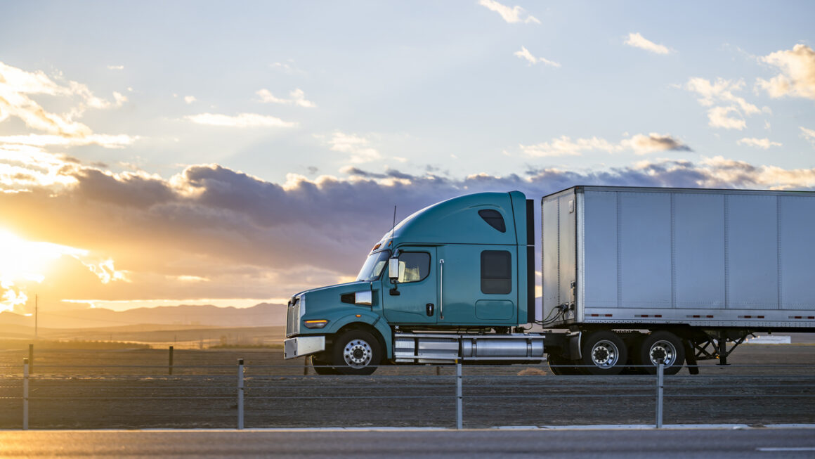Streamline green big rig bonnet semi truck transporting cargo in dry van semi trailer driving on the highway road with fields on the sides at sunset time