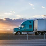Streamline green big rig bonnet semi truck transporting cargo in dry van semi trailer driving on the highway road with fields on the sides at sunset time