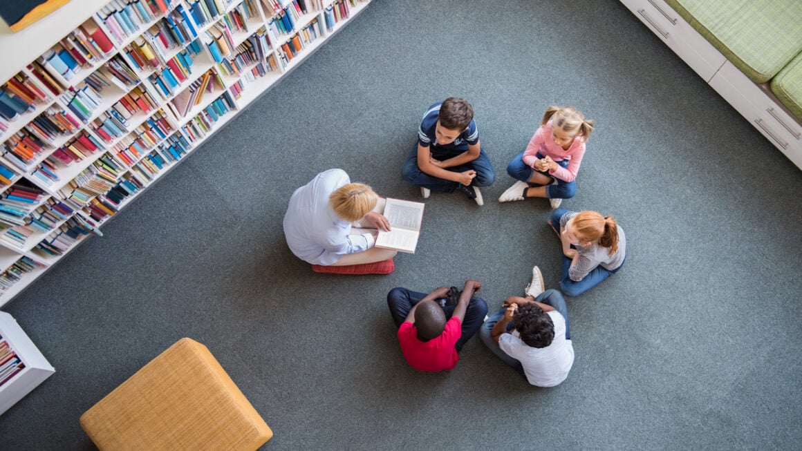 Teacher reading fairy tales to children sitting in a circle at library. Top view of librarian sitting with five multiethnic children on floor. Teacher reading book to cute girls and young boys at school.